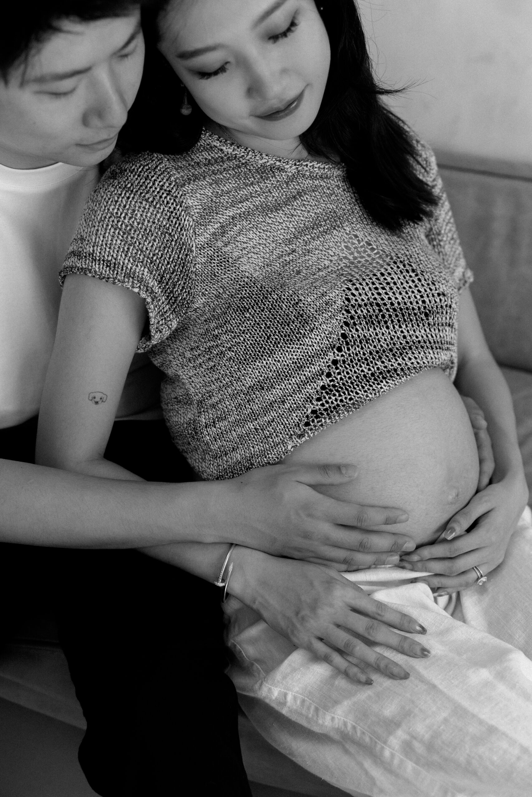 A young couple embraces on a couch with their hands on her pregnant belly.